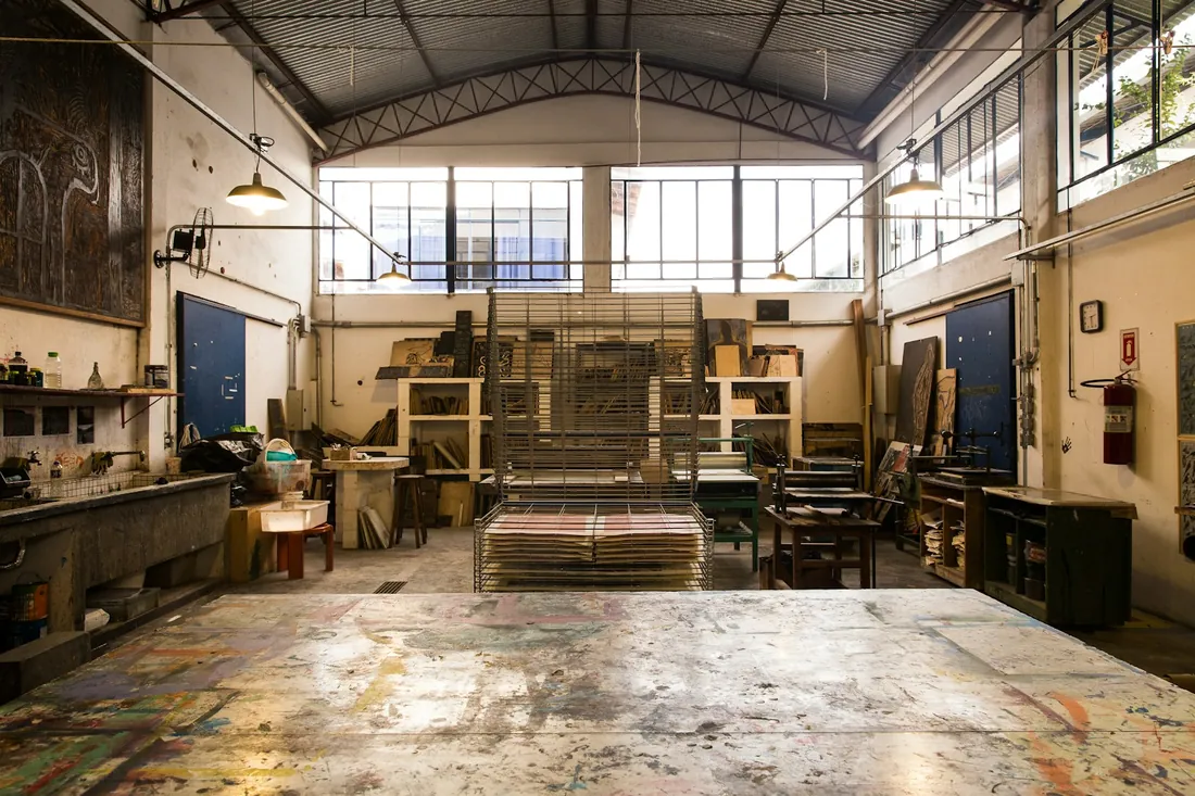 Nave de taller con mesa grande manchada de pintura, bastidores apoyados en la pared y estanterías con material de decorados
